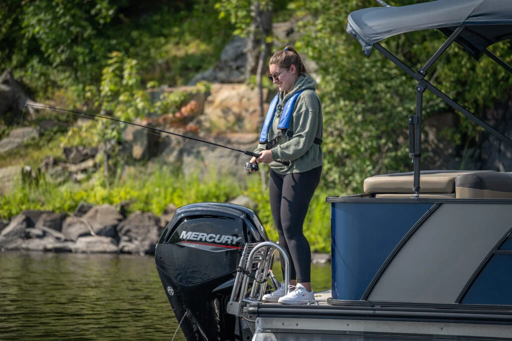 Angler wearing flotation and fishing from the stern of a Legend pontoon beside a Mercury outboard.