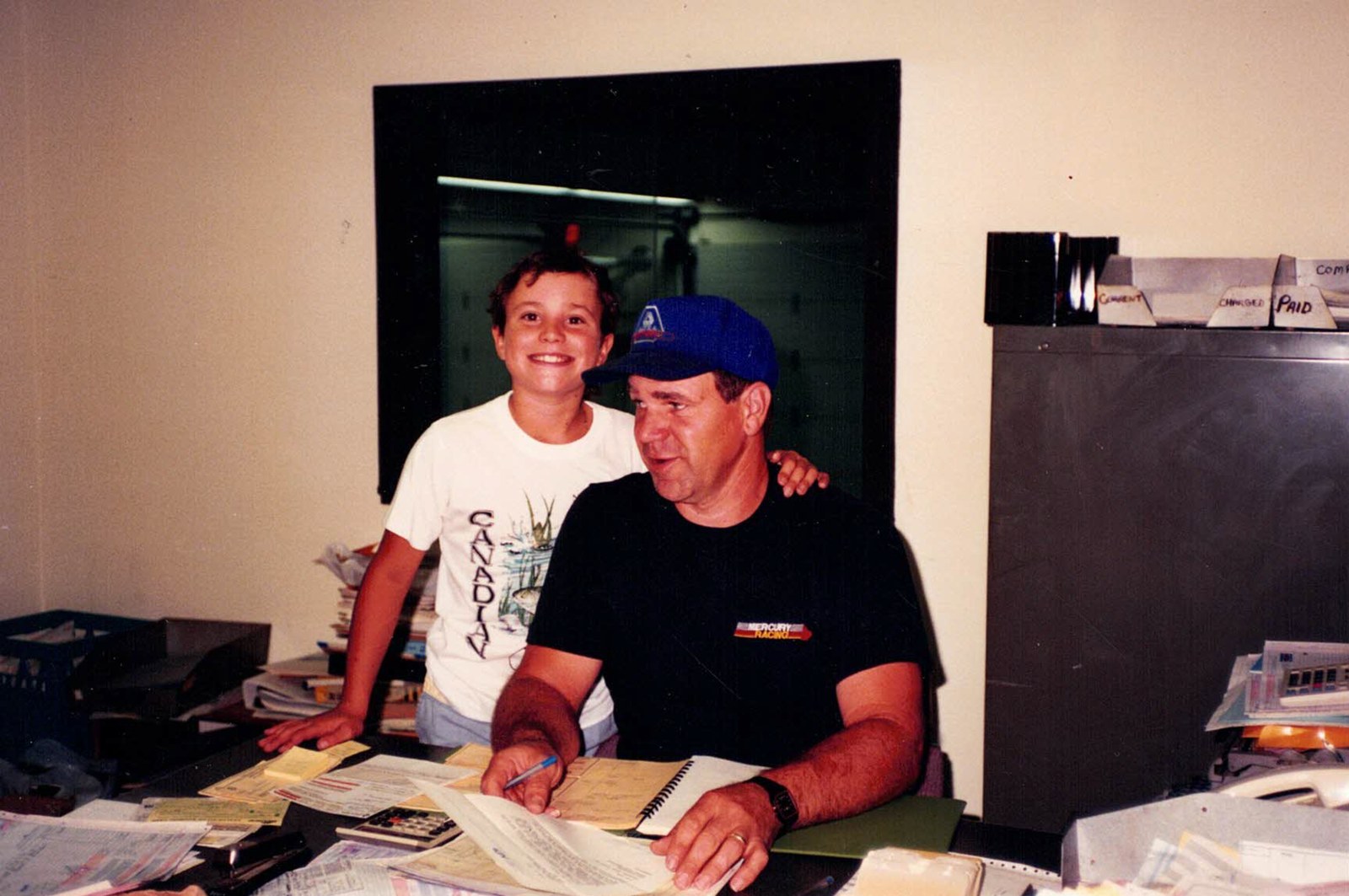 Jim Harris at his desk with a young Jay, around 1990. Paper work orders, a calculator, and a Mercury Racing t-shirt. The filing system was three tabs: current, charged, paid.